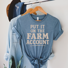 Load image into Gallery viewer, Blue t-shirt with 'Put it on the Farm Account' text hanging on a rack with denim jacket.