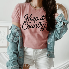 Load image into Gallery viewer, Person wearing a pink 'Keep it Country' t-shirt with a denim jacket and white shorts.