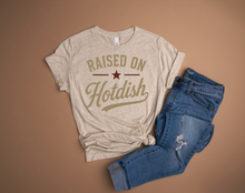 Load image into Gallery viewer, Beige t-shirt with 'Raised on Hotdish' text and blue jeans on a brown background