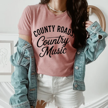 Load image into Gallery viewer, Person wearing a pink t-shirt with 'County Roads, Country Music' text, paired with a denim jacket and white shorts.