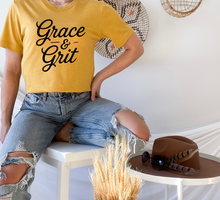 Load image into Gallery viewer, Person wearing a yellow 'Grace &amp; Grit' t-shirt sitting on a stool with a hat and sunglasses on a table.