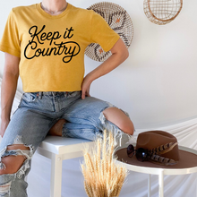 Load image into Gallery viewer, Person wearing a yellow 'Keep it Country' t-shirt sitting on a chair with a hat and sunglasses on a small table.