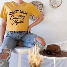 Load image into Gallery viewer, Person wearing a yellow t-shirt with 'County Roads, Country Music' text, sitting on a chair with a hat and sunglasses.
