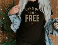 Load image into Gallery viewer, Person wearing a black t-shirt with 'Land of the Free' text, standing indoors with plants and a textured wall.