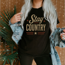 Load image into Gallery viewer, Person wearing a black t-shirt with 'Stay Country' text, standing indoors with plants and a textured wall in the background.