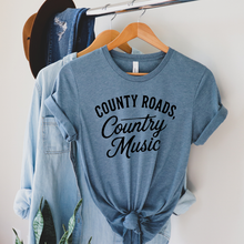 Load image into Gallery viewer, Blue t-shirt with 'County Roads, Country Music' text on a hanger with denim jacket and hat.