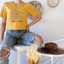Load image into Gallery viewer, Person wearing a yellow 'Stay Country' t-shirt sitting on a chair with a hat and sunglasses on a small table.