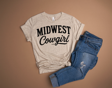 Load image into Gallery viewer, Beige t-shirt with 'Midwest Cowgirl' text and blue jeans on a brown background